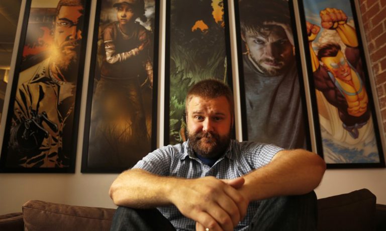 Robert Kirkman sentado em um sofá com alguns quadros de seus projetos ao fundo