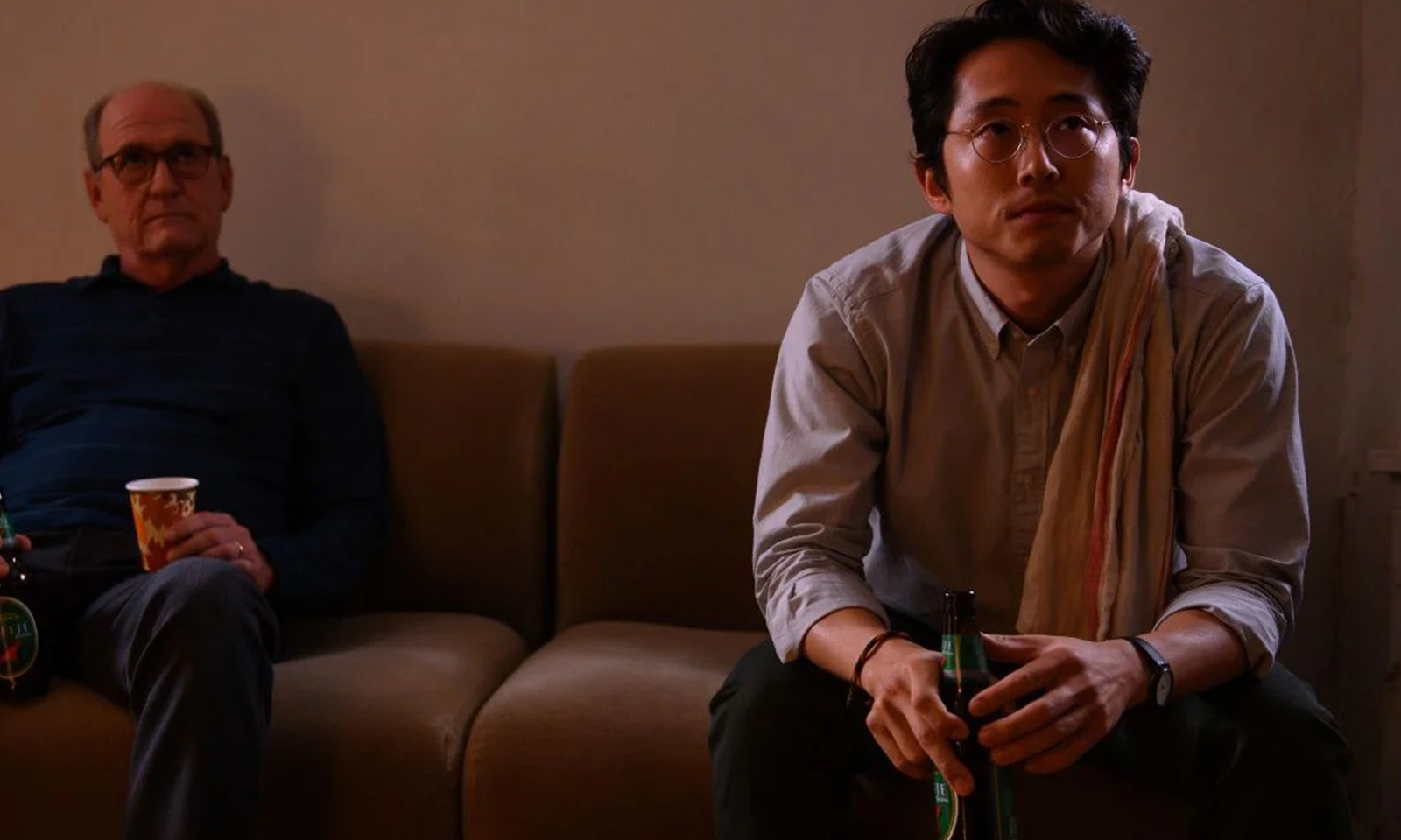 Steven Yeun como Richard sentado no sofá tomando uma cerveja em imagem do filme The Humans.