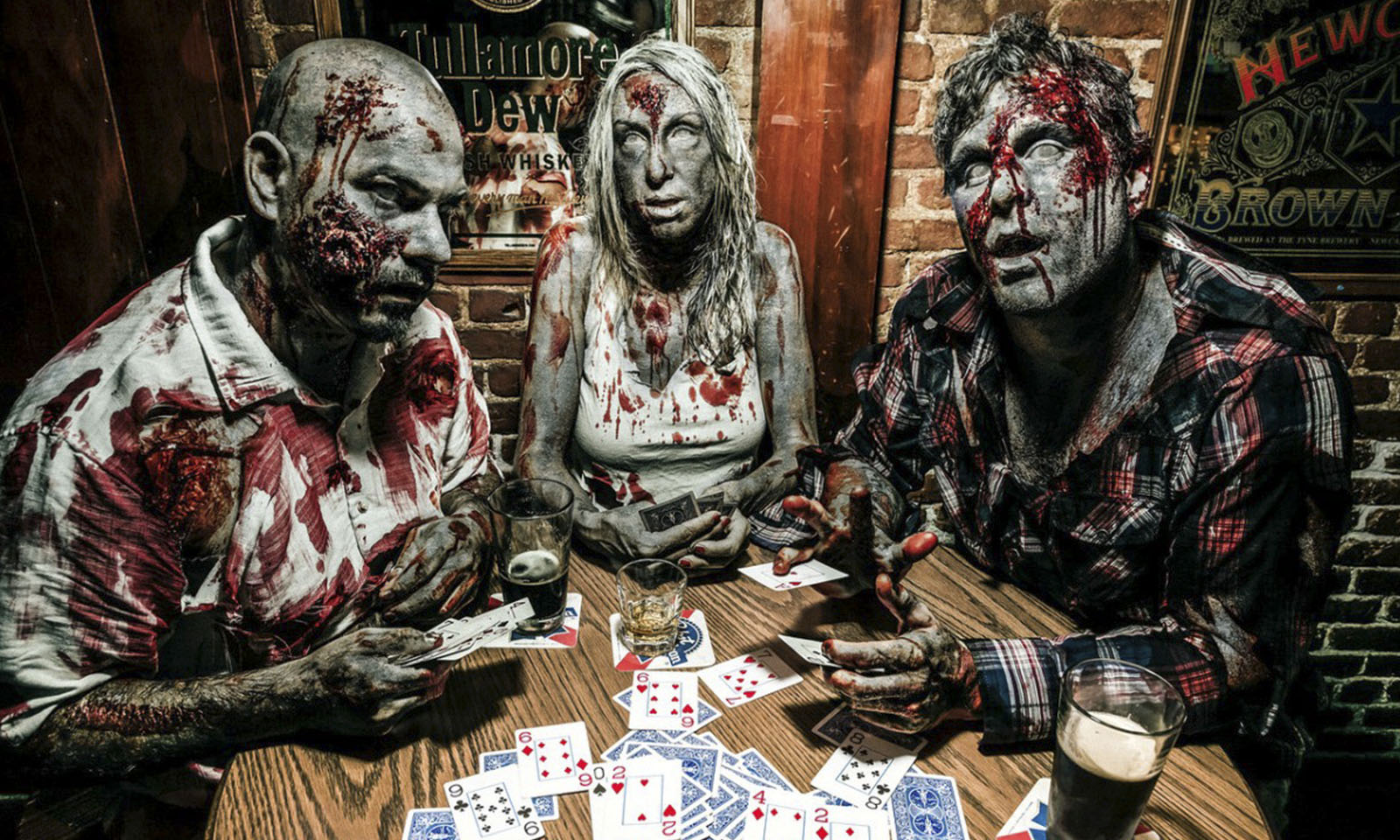 Três pessoas com maquiagem de zumbis jogando poker em uma mesa cheia de cartas.