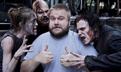 Robert Kirkman fazendo joinha ao lado de 3 zumbis da 1ª temporada de The Walking Dead.