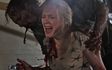 Cena intensa de ataque: uma mulher (Emma Bell, como Amy) loira grita de dor enquanto é mordida no ombro por um homem com aparência ferida e ensanguentada. Há sangue visível escorrendo pelo pescoço e pela roupa dela, que está rasgada. O agressor está atrás dela, segurando-a com força. O ambiente é escuro e caótico, reforçando a sensação de perigo e desespero. Em cena na 1ª temporada de The Walking Dead