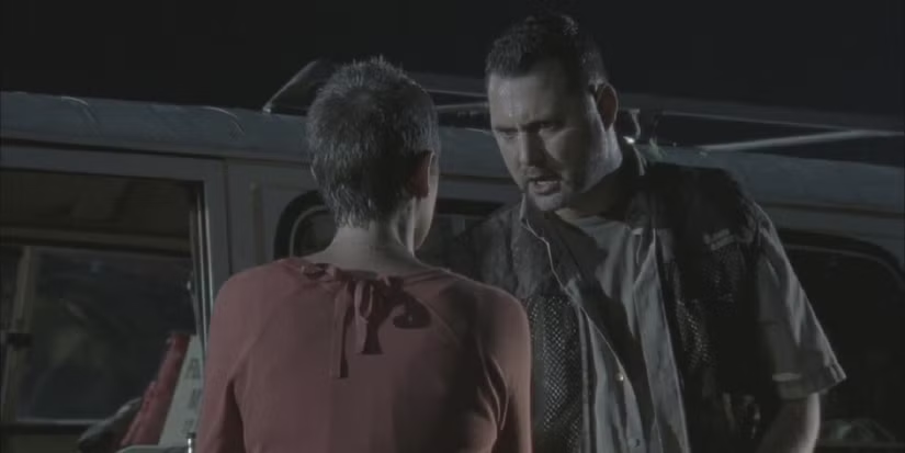 Cena noturna: um homem de expressão agressiva encara uma mulher de costas, que usa uma blusa vermelha. Eles estão próximos a um veículo estacionado, sob iluminação baixa. A postura do homem sugere confronto, enquanto a mulher parece tensa e retraída, reforçando um clima de conflito e intimidação.