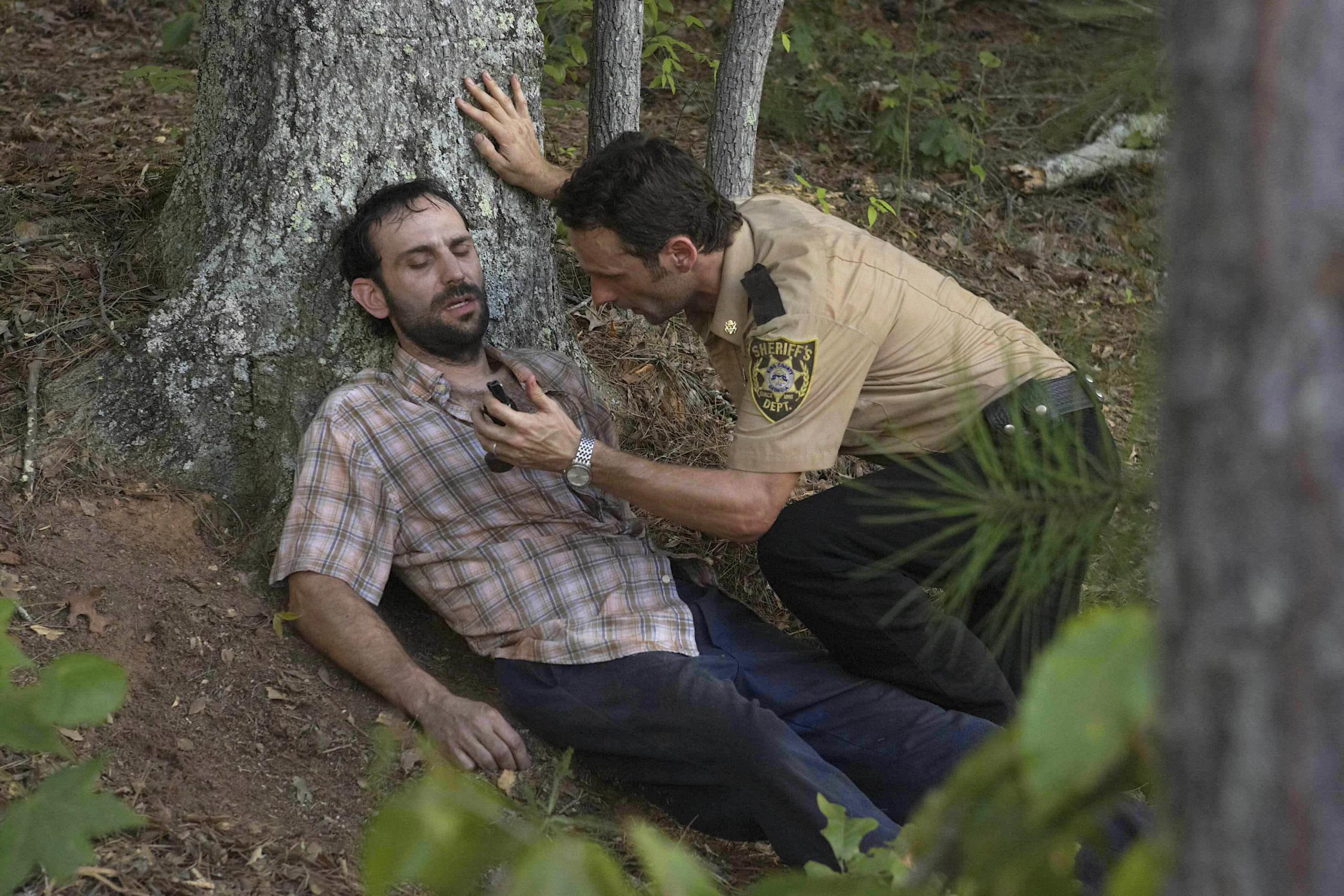 Em uma área de mata, um homem (Andrew Rothenberg, como Jim) está caído ao pé de uma árvore, com aparência fraca e ferida, olhos semiabertos e corpo apoiado no tronco. Ao lado dele, um policial (Andrew Lincoln como Rick) com uniforme de xerife se inclina, segurando-o pelo peito enquanto verifica seu estado com expressão tensa e preocupada. O chão está coberto por folhas secas e vegetação, reforçando o clima de urgência e sobrevivência em meio à natureza. mortes da primeira temporada de The Walking Dead