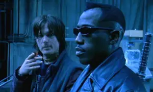 Dois homens (Norman Reedus e Weslwy Snipes) estão em um ambiente industrial com iluminação azulada e fria. Em primeiro plano, um homem negro usa óculos escuros e um casaco de couro, com expressão séria, olhando para frente. Ao fundo, um homem de cabelos médios e bagunçados segura um cigarro próximo ao rosto, vestindo roupas escuras e um colar no pescoço, observando o outro. O cenário inclui paredes metálicas e estruturas industriais, reforçando uma atmosfera tecnológica e sombria.