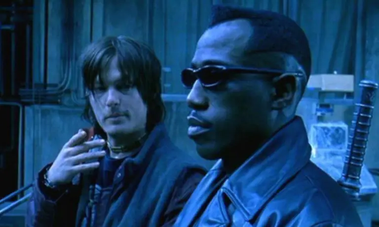 Dois homens (Norman Reedus e Weslwy Snipes) estão em um ambiente industrial com iluminação azulada e fria. Em primeiro plano, um homem negro usa óculos escuros e um casaco de couro, com expressão séria, olhando para frente. Ao fundo, um homem de cabelos médios e bagunçados segura um cigarro próximo ao rosto, vestindo roupas escuras e um colar no pescoço, observando o outro. O cenário inclui paredes metálicas e estruturas industriais, reforçando uma atmosfera tecnológica e sombria.