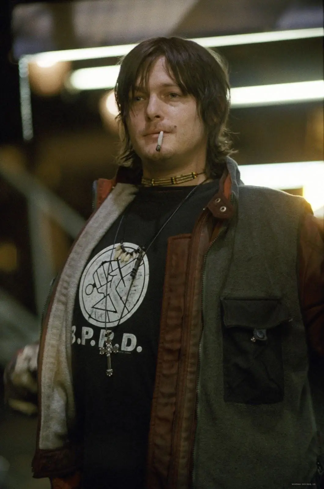 Ator Norman Reedus, com os cabelos escuros e bagunçados encara a câmera com um leve sorriso, com um cigarro entre os lábios. Ele usa um colar estilo choker e roupas escuras, com aparência urbana. A cena acontece à noite, em um ambiente externo iluminado por luzes artificiais, com estruturas metálicas ao fundo, criando uma atmosfera fria e levemente industrial, cena do filme Blade II: O Caçador de Vampiros.