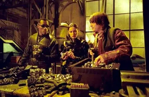 Três pessoas Wesley Snipes (como Blade), Leonor Varela (como Nyssa) e Norman Reedus (como Scud), estão reunidos em um ambiente com aparência de laboratório ou oficina industrial, iluminado por luz amarelada. Sobre a mesa à frente, há várias armas e equipamentos organizados. À esquerda Wesley Snipes (como Blade), um homem de óculos escuros e roupa preta observa com postura séria. No centro Leonor Varela (como Nyssa), uma mulher com roupas táticas examina um objeto com atenção. À direita Norman Reedus (como Scud), um homem de cabelo médio, vestindo jaqueta escura, mexe em uma maleta aberta cheia de itens, como se estivesse preparando ou ajustando equipamentos. O cenário ao fundo tem estruturas metálicas e janelas grandes, reforçando o clima tecnológico e sombrio.