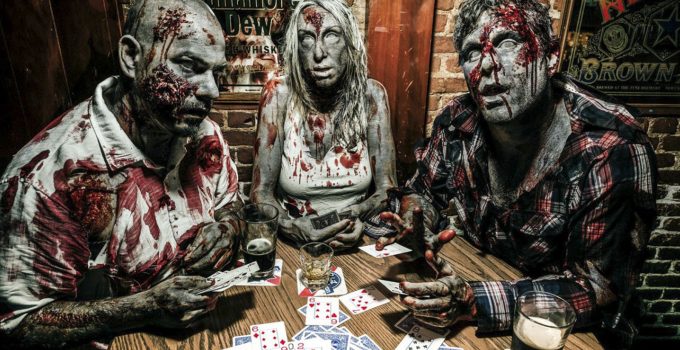 Três pessoas com maquiagem de zumbis jogando poker em uma mesa cheia de cartas.