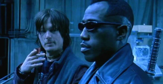 Dois homens (Norman Reedus e Weslwy Snipes) estão em um ambiente industrial com iluminação azulada e fria. Em primeiro plano, um homem negro usa óculos escuros e um casaco de couro, com expressão séria, olhando para frente. Ao fundo, um homem de cabelos médios e bagunçados segura um cigarro próximo ao rosto, vestindo roupas escuras e um colar no pescoço, observando o outro. O cenário inclui paredes metálicas e estruturas industriais, reforçando uma atmosfera tecnológica e sombria.