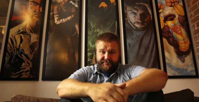 Robert Kirkman sentado em um sofá com alguns quadros de seus projetos ao fundo